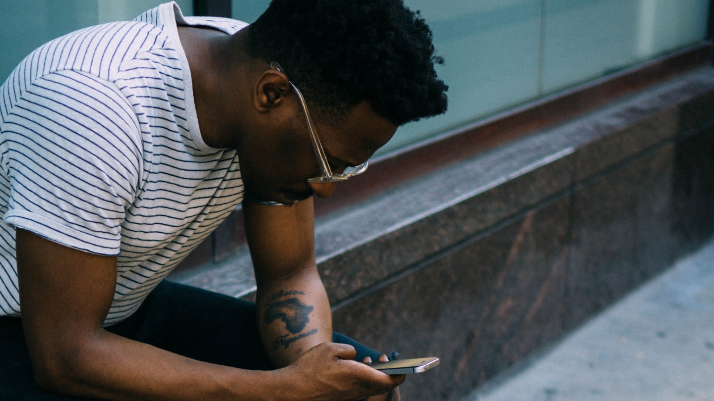 A Black man in glasses and a t-shirt sitting on a marble wall, looking at a phone.