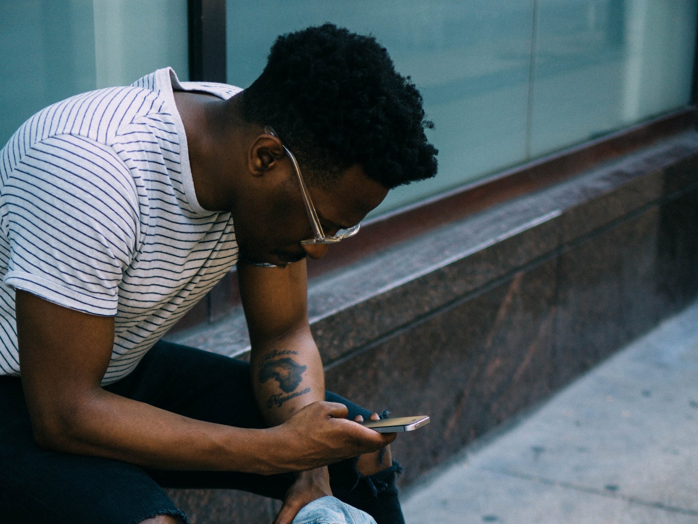A Black man in glasses and a t-shirt sitting on a marble wall, looking at a phone.