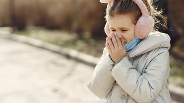 A young girl stands outside in a winter coat, pink earmuffs, and a blue face mask pulled down over her chin as she sneezes into her hands.