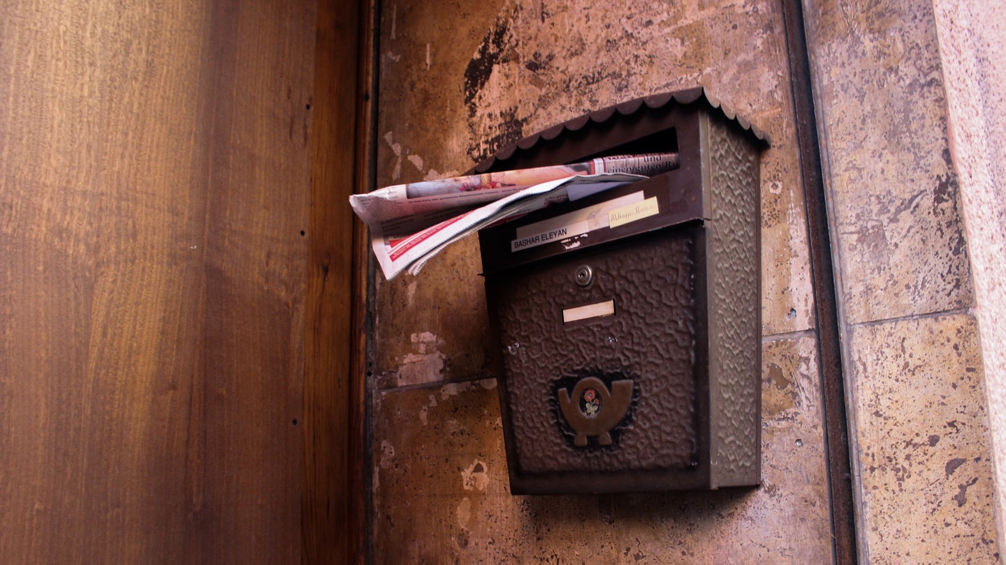 An old metal mailbox mounted on a wall and overflowing with junk mail.
