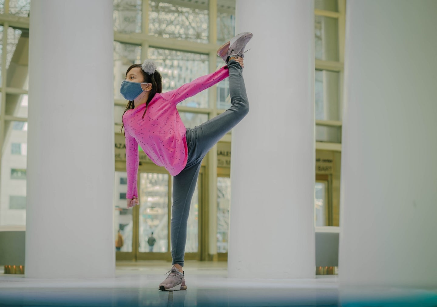 Child in a pink shirt and hair bow wearing a mask and holding a dance pose inside an atrium