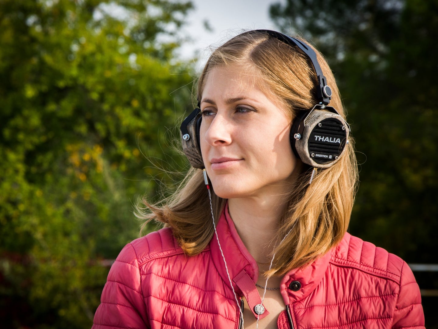 A woman in a pink jacket outside near some trees, listening to music on a pair of Thalia headphones.