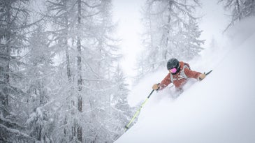 person in an orange ski jacket and black ski helmet, skiing down a powdery mountain with snow