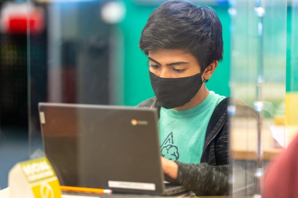 Student in black mask and green shirt on a laptop behind plexiglass
