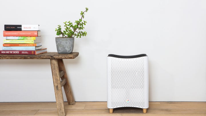 Mila air purifier, one of the best air purifiers, on a wood floor next to a wooden bench with a plant and books on it