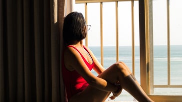 A woman in a red bathing suit sitting on the floor by a sliding glass door, staring at the ocean outside.