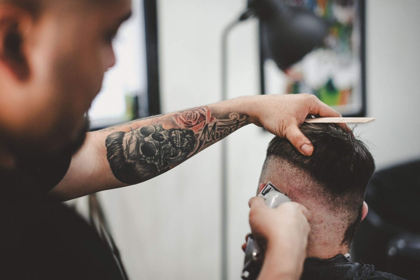 man with tattooed arms using clippers to cut another man's hair