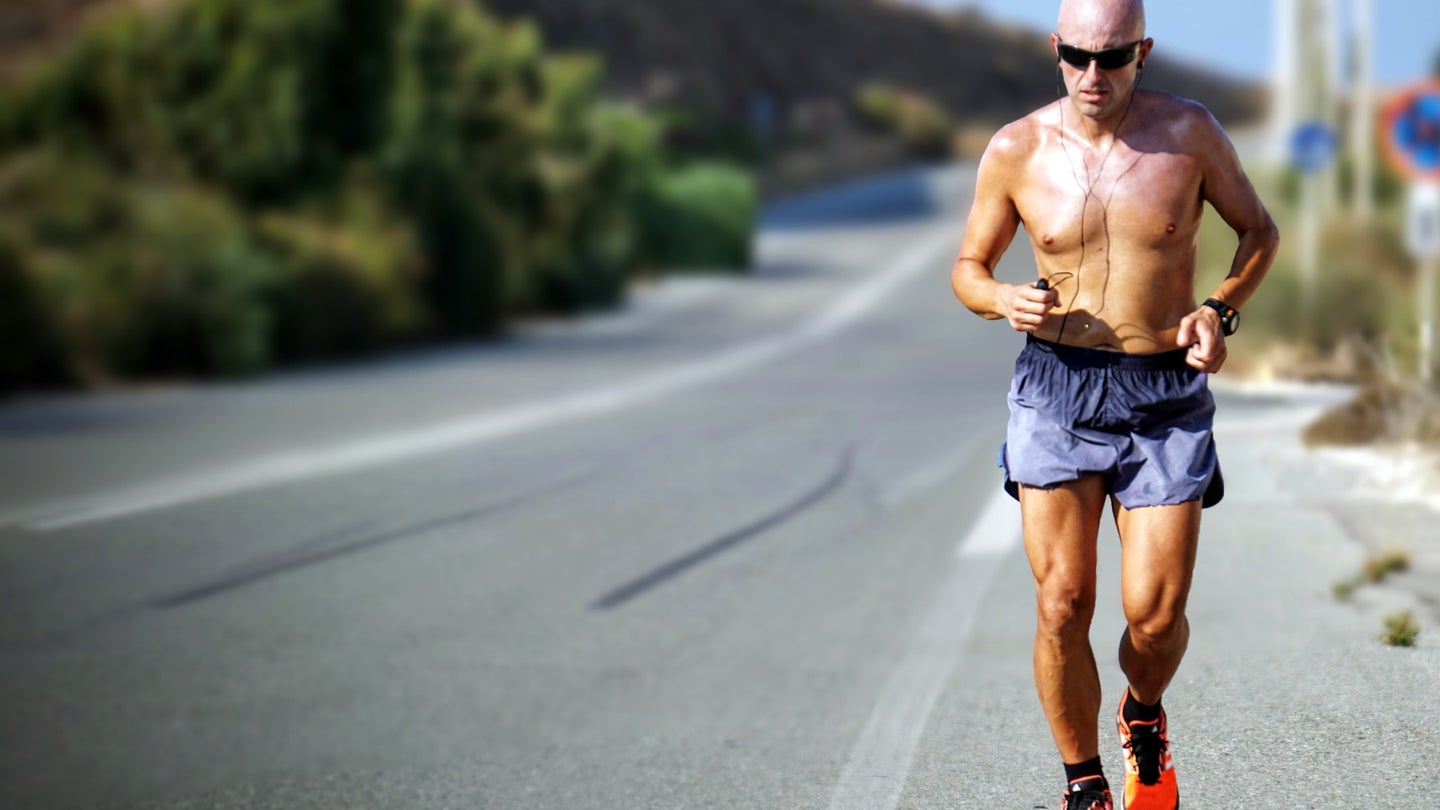 A man running shirtless on the side of a road in hot weather.
