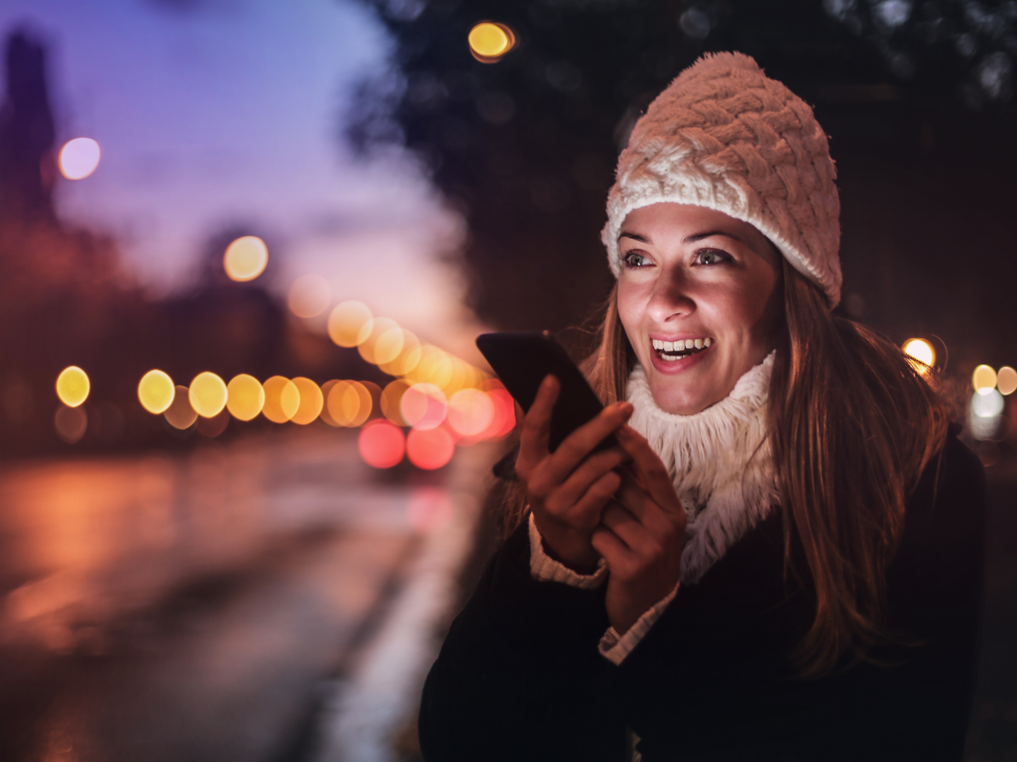 A woman wearing a white knit cap, a white sweater, and a jacket, talking into a phone at night by a street.