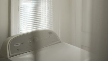 A white clothes dryer behind a white curtain, in front of a tall window with sunlight shining through.
