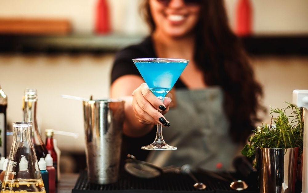 A smiley girl that has in her hands a sky color cocktail beside a silver shaker cocktail glass.