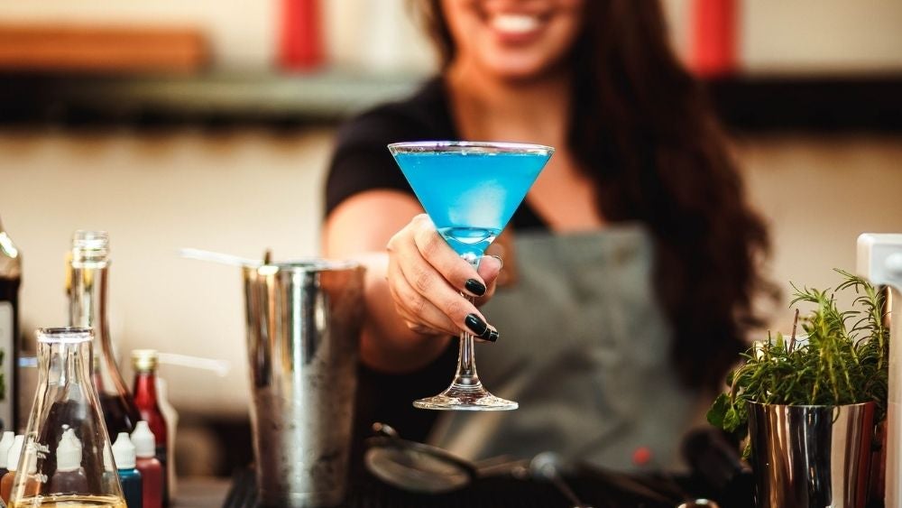 A smiley girl that has in her hands a sky color cocktail beside a silver shaker cocktail glass.