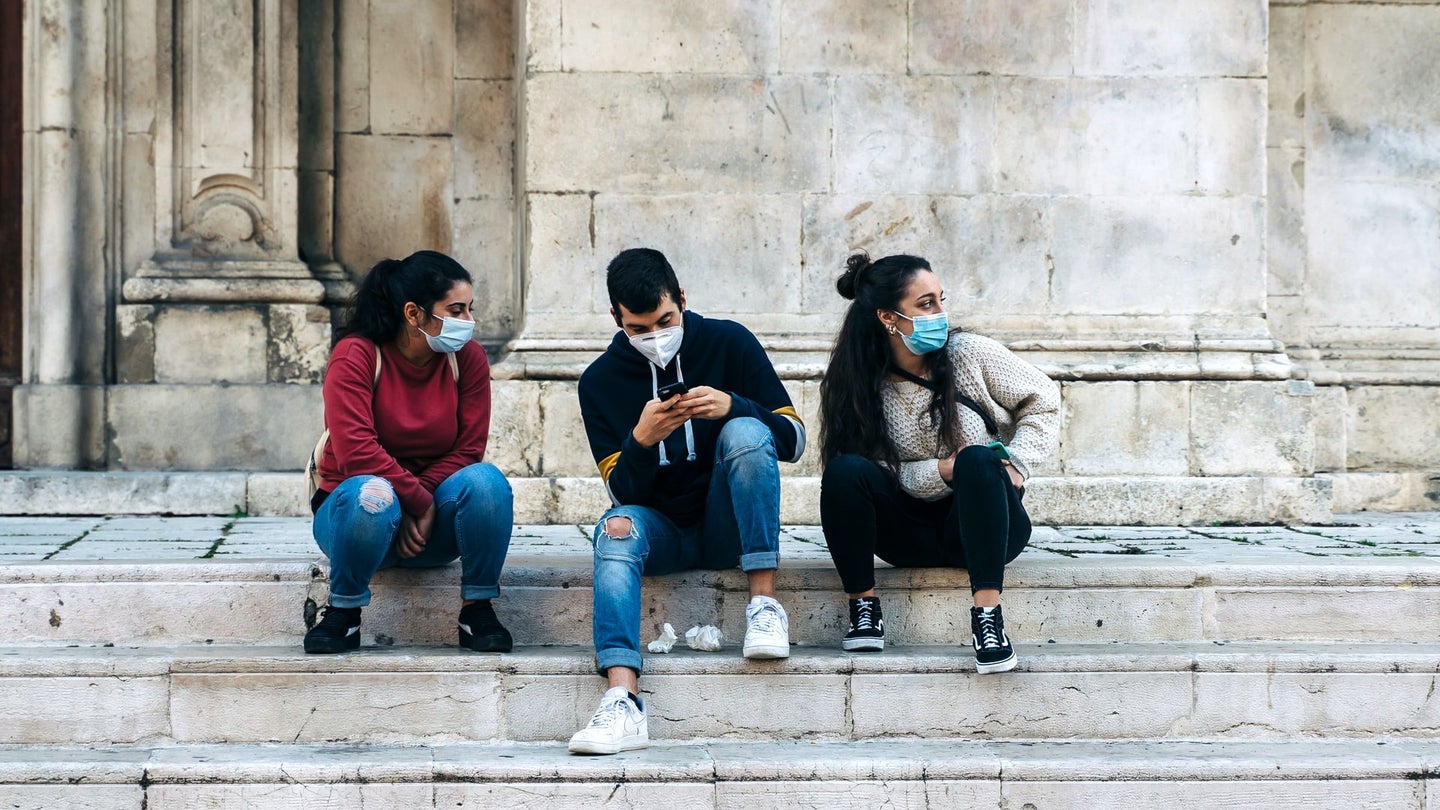 Teens in masks sit on marble stairs.