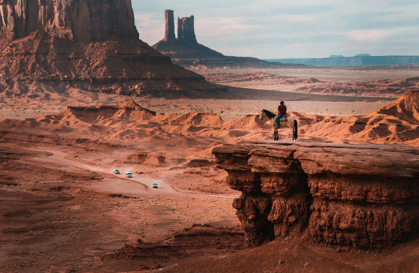 monument valley with person on horse in foreground cars in background