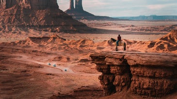 monument valley with person on horse in foreground cars in background