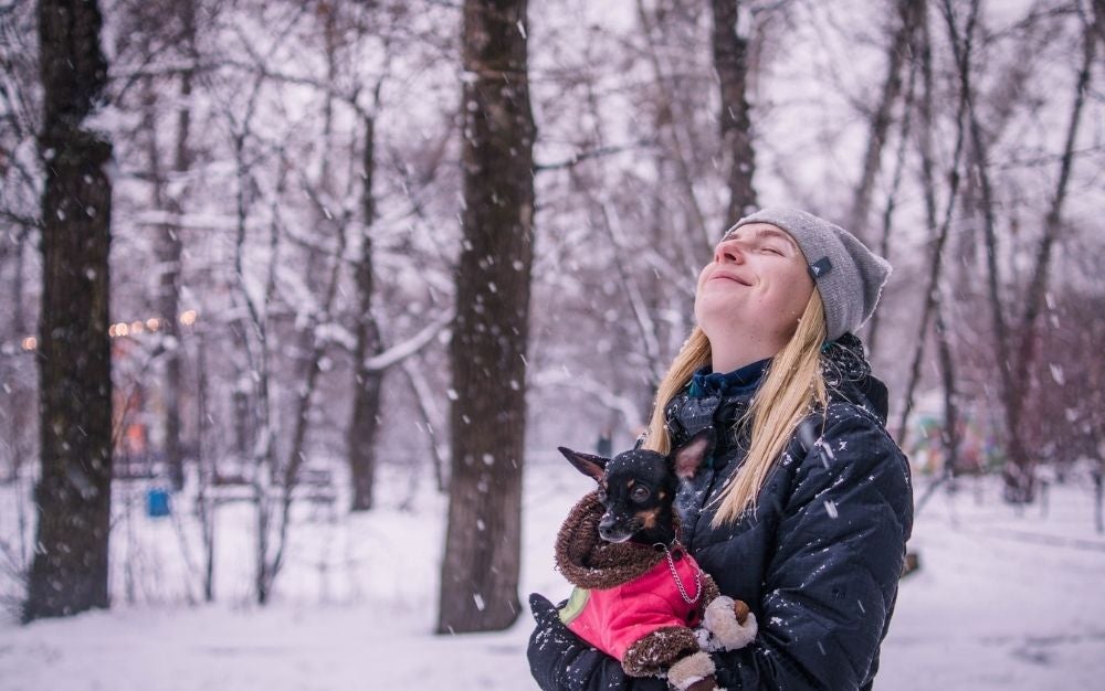 A blonde girl wearing a winter jacket and a dog in her hand as she closes her eyes and is enjoying the snow.