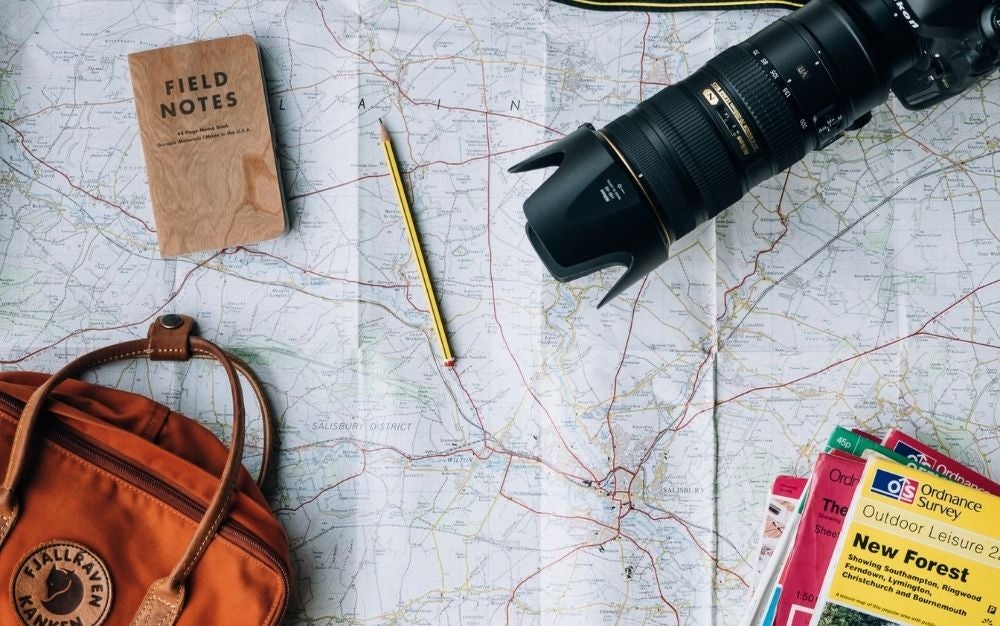 A map with the directions of the roads and over it a pencil, a professional camera, a notebook, and an orange bag.