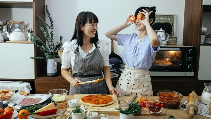 Two woman laughing with each other while cooking pizza in the kitchen.