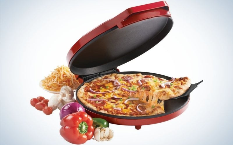 A red metal pizza oven with a pizza into it and some vegetables on his side.
