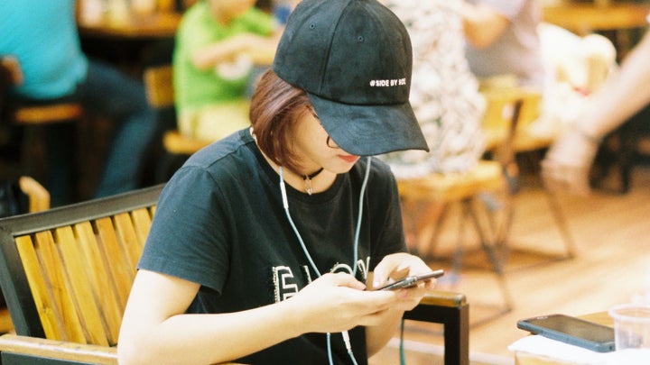 A person sitting at a coffee shop, using a phone while wearing headphones and a cap.