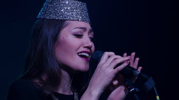 Person with long black hair and in silver sequin hat at microphone