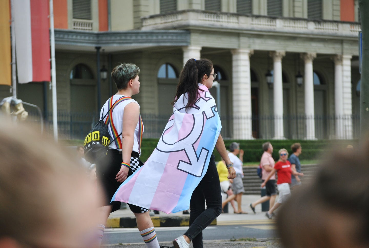 Teen with trans flag over shoulders walking with friend in rainbow pride suspenders