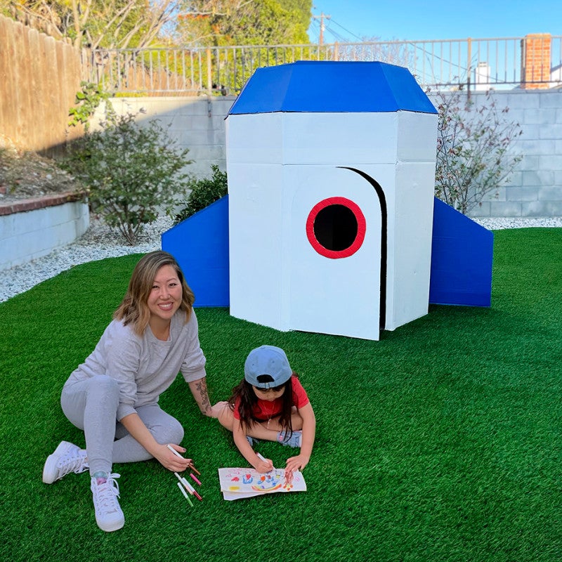 Mom and kid in front of a cardboard rocket with white, blue, and red pain in a fenced-in yard