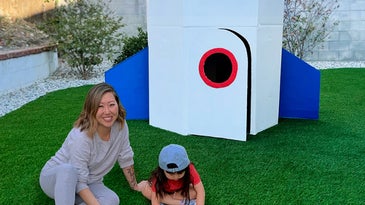 Mom and kid in front of a cardboard rocket with white, blue, and red pain in a fenced-in yard