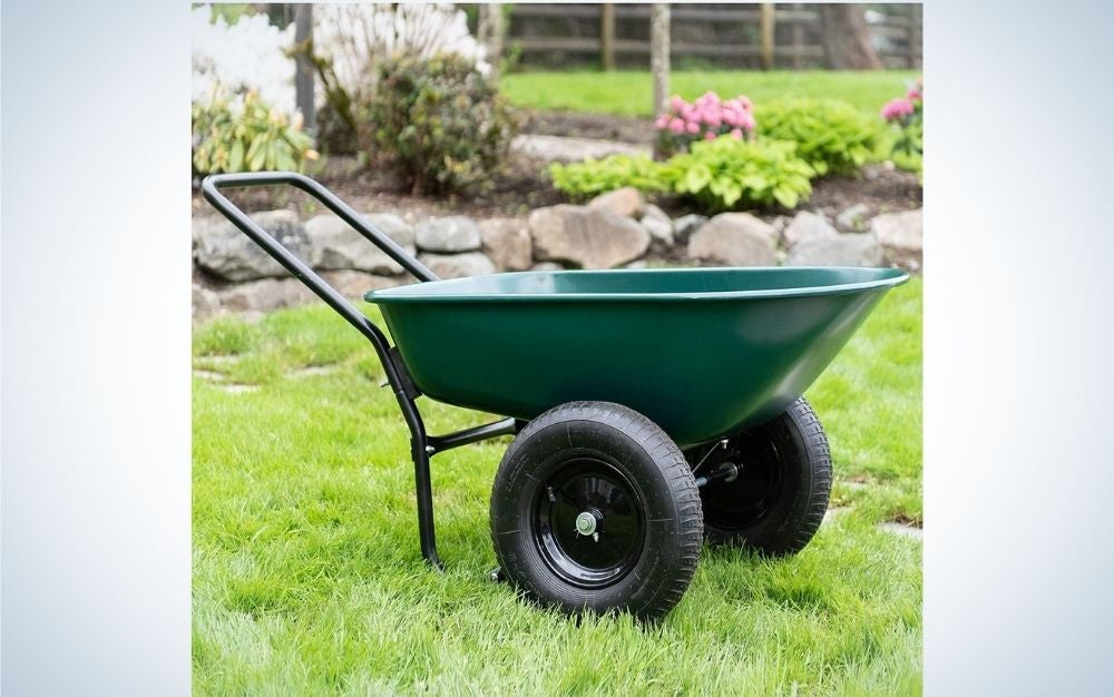 A small wheelbarrow in a green color with two black wheels standing over a grassy ground and flowers.