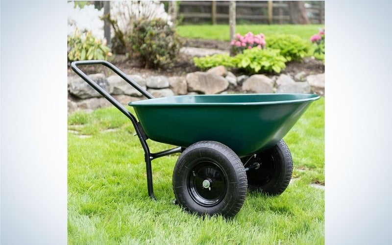 A small wheelbarrow in a green color with two black wheels standing over a grassy ground and flowers.