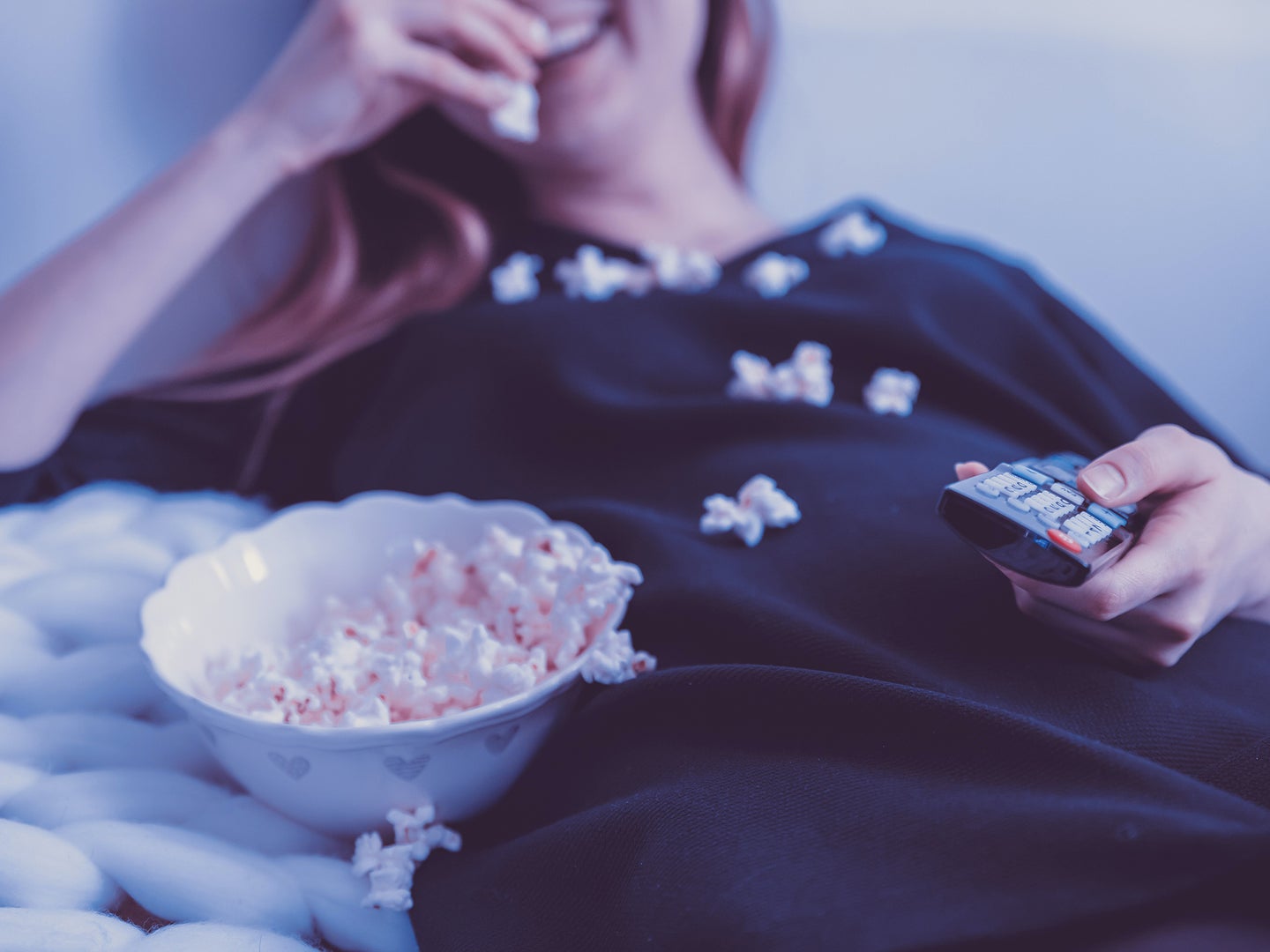 A woman wearing a baggy black shirt lounging on a couch, eating a bowl of popcorn, while holding a TV remote with popcorn all over her chest.