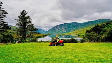 A man with a hat riding a four wheel lawn mower with black and red color into a green field all of grass in front of a lake.