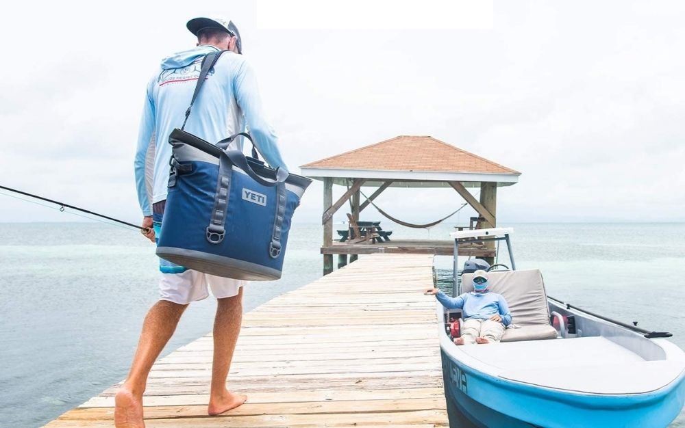 A man with shorts and with a hat and a big YETI bag walking into a wooden place through the sea.