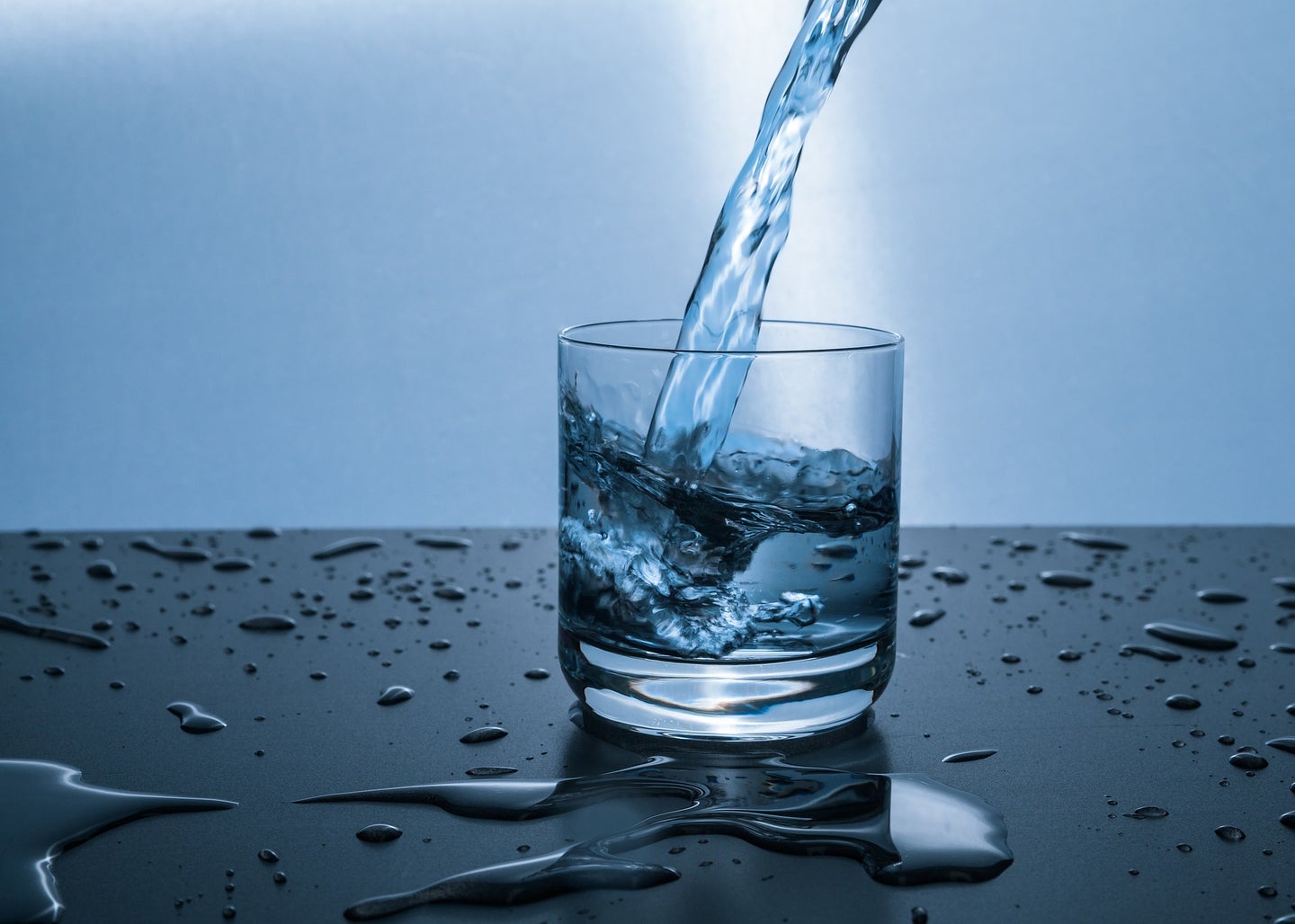 Water is poured into a short, clear glass from above against a blue background.