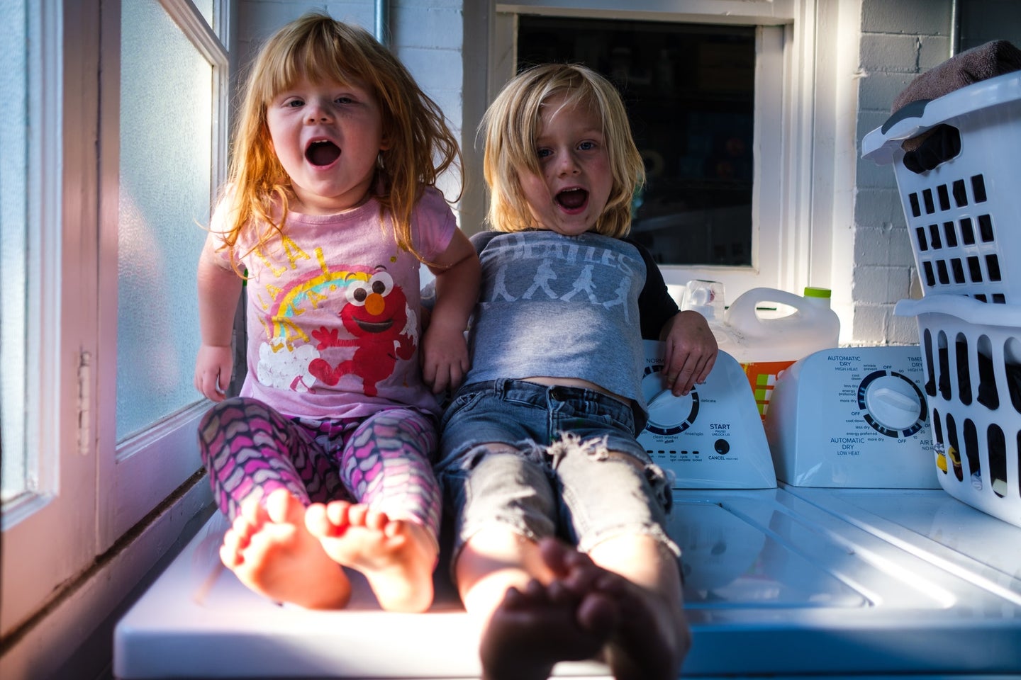 Two kids with blond and red hair sitting on a washer in a laundry room