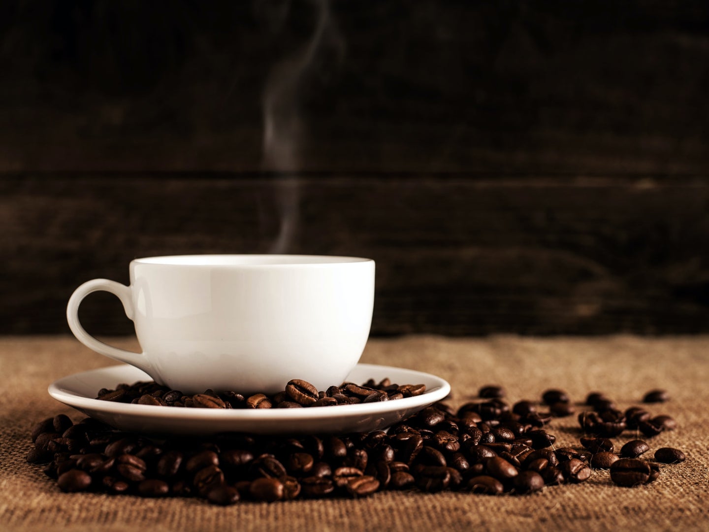 A white mug full of hot coffee on a white saucer, surrounded by roasted coffee beans.