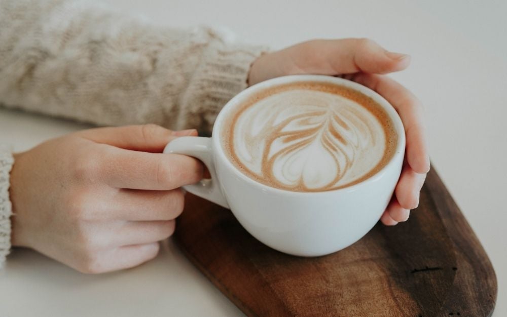 A girl with a beige sweater holding in her hands a cup of coffee with a leave formed on it.