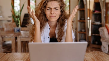 A woman with curly, dirty blonde hair sitting in front of a gray laptop with her hands in the air in an act of confusion or frustration.