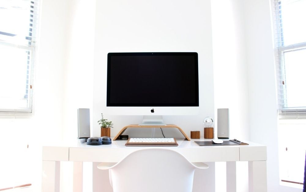 A white Imac with off screen which stands on a white stand monitor and a keyboard and full of other computer accessories.