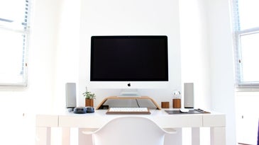 A white Imac with off screen which stands on a white stand monitor and a keyboard and full of other computer accessories.