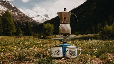 Moka pot on a burner in front of a snowy mountain with two enamel cups