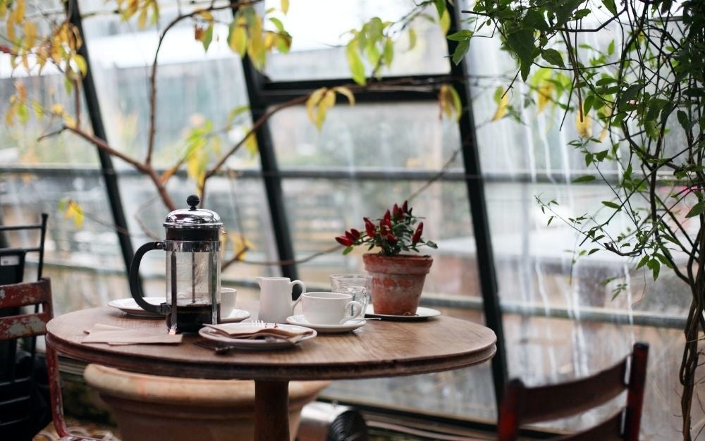 A table on a glass balcony, the table is wooden and has white coffee cups on top and a kettle to keep the coffee warm.
