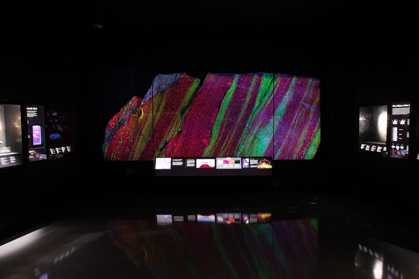 A large piece of granite rock glows in bright green and crimson red stripes behind glass and against the dark background of a museum exhibit. A few illegible, backlit display signs surround the large rock.