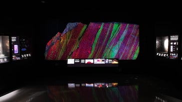 A large piece of granite rock glows in bright green and crimson red stripes behind glass and against the dark background of a museum exhibit. A few illegible, backlit display signs surround the large rock.