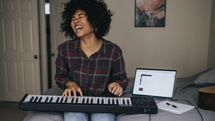 A person sitting on a bed, enjoying playing a keyboard that's hooked up to a laptop.