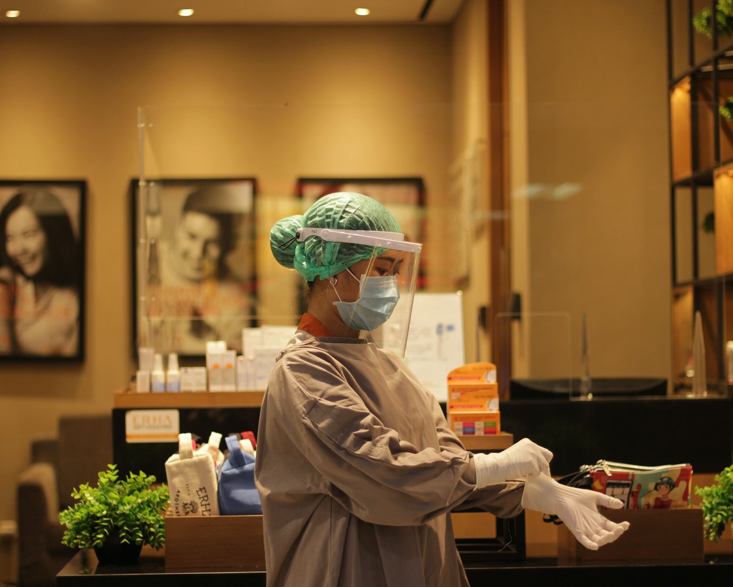 A person wearing a surgical hair net, face mask, face shield and protective surgical gown pulls on surgical gloves while standing in a health clinic.