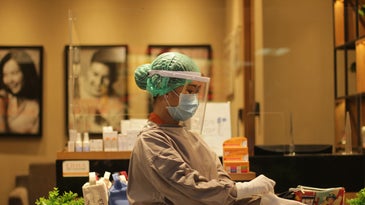 A person wearing a surgical hair net, face mask, face shield and protective surgical gown pulls on surgical gloves while standing in a health clinic.