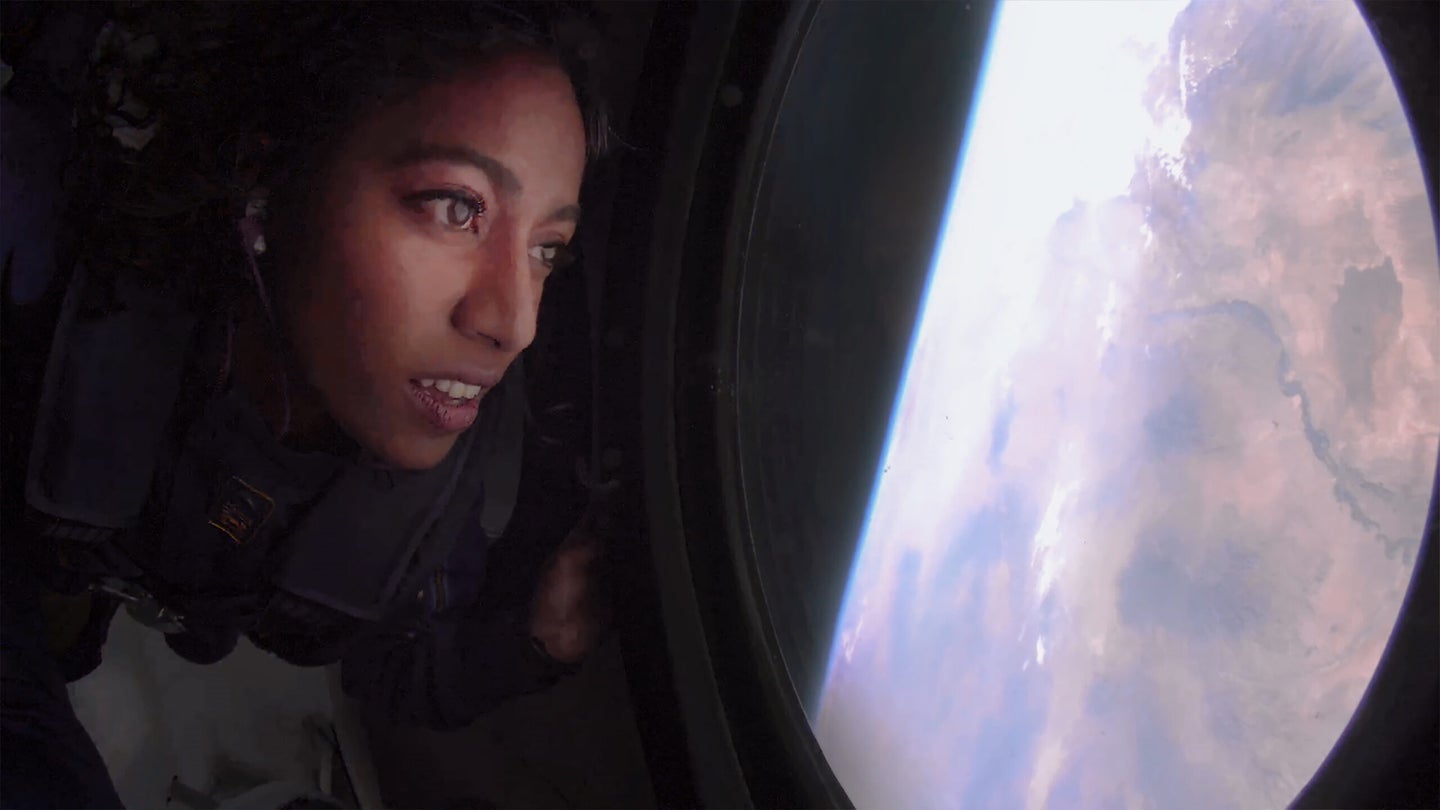 A woman's face partially in shadow, aboard a suborbital, looks out a window back at Earth.