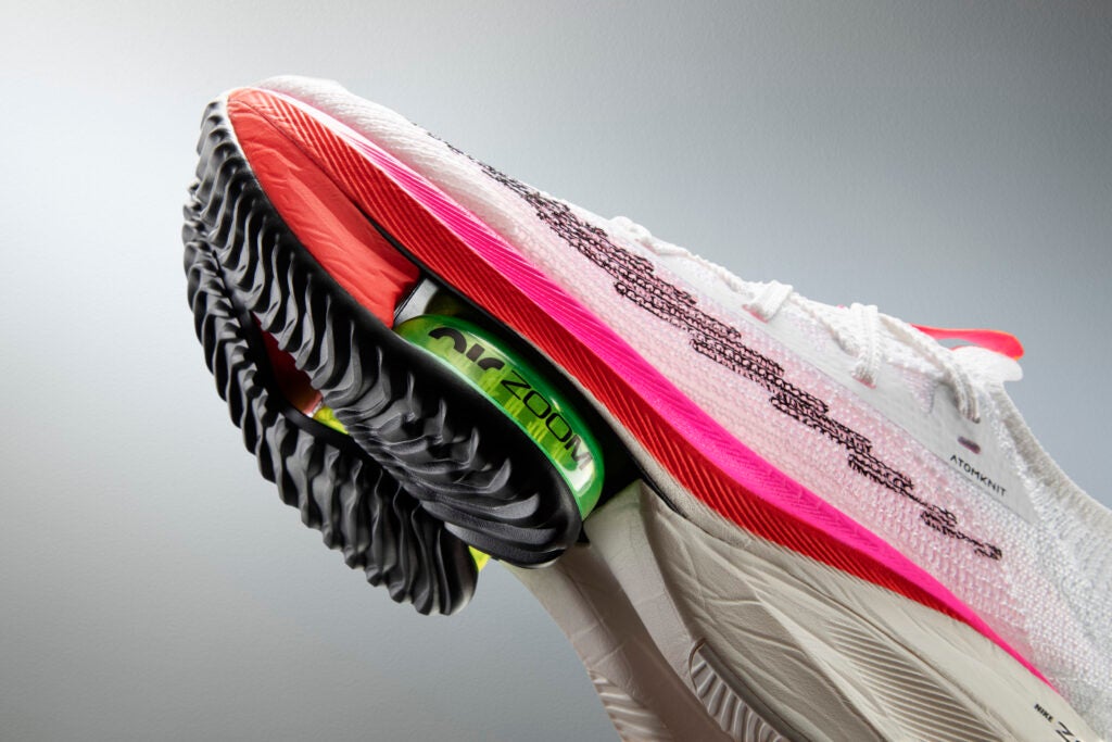 The front half of a mostly white running shoe with grey, red, neon pink, and lime green accents is pictured at a diagonal angle against a grey background.