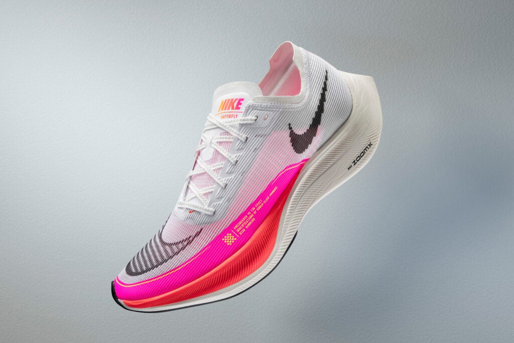 A mostly white running shoe with neon pink and orange accents floats at a diagonal angle against a grey background. The Nike swoosh is prominently visible in grey on the shoe's side. The word "Nike" is written in red on the tongue.